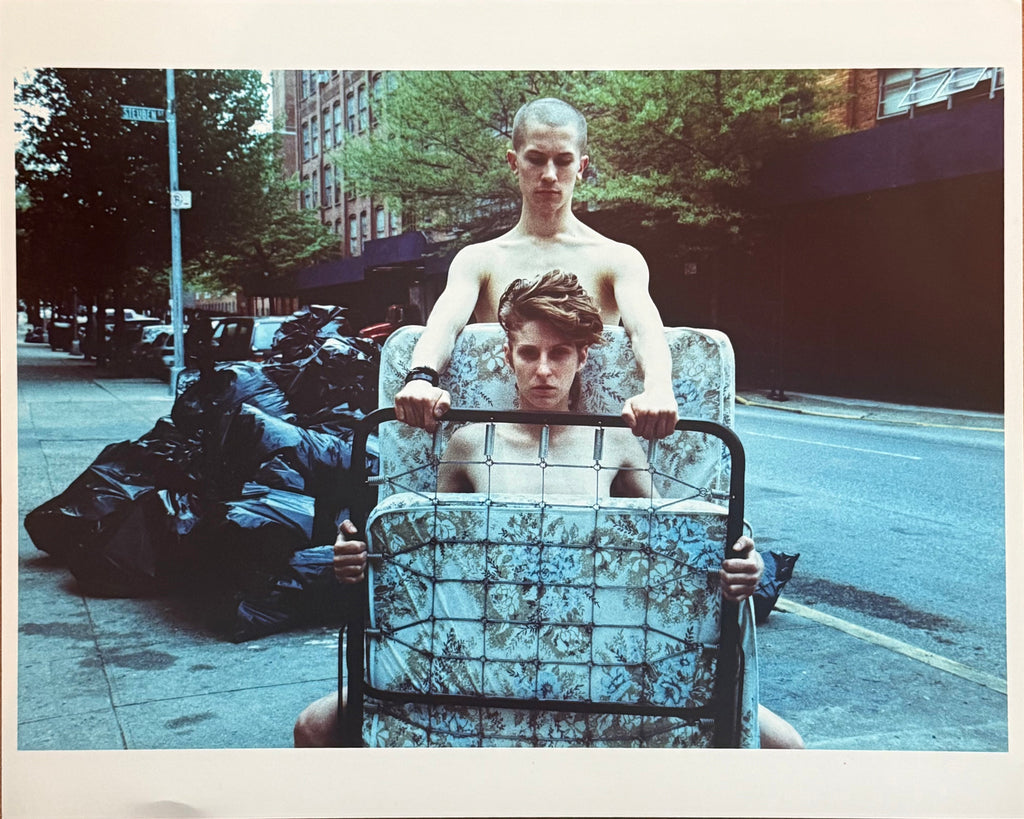 Colour photograph of a young man wheeling a mattress and bed frame on a hand truck along a New York sidewalk